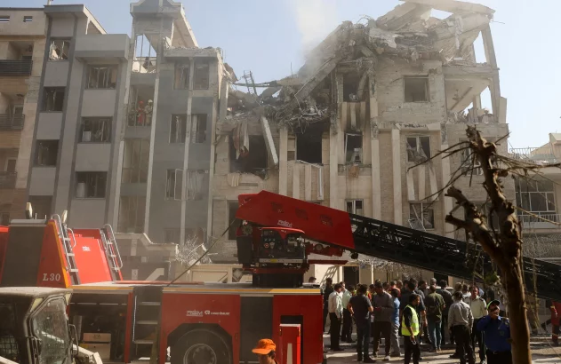 Edifício parcialmente desabado, fumaça. Caminhão de bombeiros com escada estendida. Equipes de resgate e multidão.