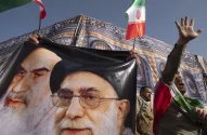 Pessoas seguram banner de Khomeini e Khamenei com bandeiras do Irã; um participante levanta o braço.