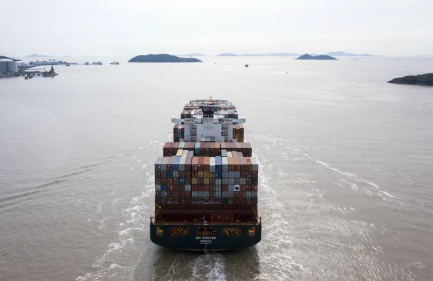 Navio porta-contêineres ZIM CHICAGO navegando, visto de popa, com contêineres coloridos. Ilhas e porto ao fundo.