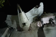 Cauda e asa de avião embutidas em prédio destruído à noite. Equipamento de resgate/demolição visível nos escombros.