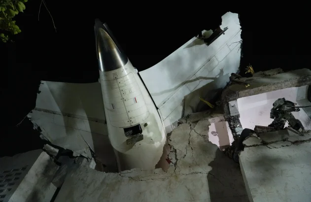 Cauda e asa de avião embutidas em prédio destruído à noite. Equipamento de resgate/demolição visível nos escombros.