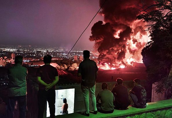 Pessoas observam grande incêndio noturno com chamas e fumaça sobre uma cidade.