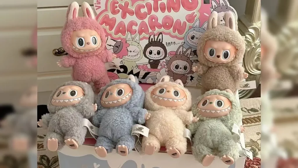Seis bonecos de pelúcia Labubu com rosto de monstro, orelhas de coelho e dentes, em várias cores, expostos em frente a uma caixa.