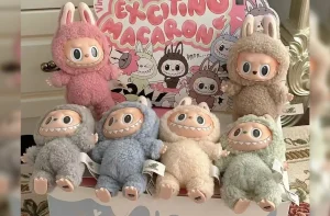 Seis bonecos de pelúcia Labubu com rosto de monstro, orelhas de coelho e dentes, em várias cores, expostos em frente a uma caixa.