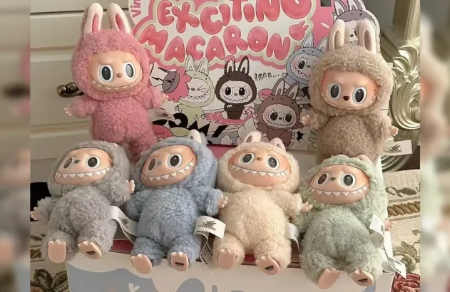Seis bonecos de pelúcia Labubu com rosto de monstro, orelhas de coelho e dentes, em várias cores, expostos em frente a uma caixa.