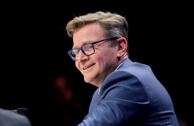 Homem sorrindo de óculos e terno azul, olhando para o lado esquerdo em um fundo escuro.