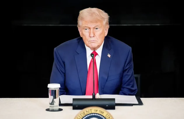 Donald Trump está de terno azul e grvata vermelho sentado com uma mesa em frente olhando para direita