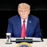 Donald Trump está de terno azul e grvata vermelho sentado com uma mesa em frente olhando para direita