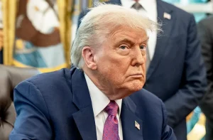 Donald Trump, de terno azul com pin da bandeira, sentado em um escritório. Ele tem um olhar sério e pensativo.