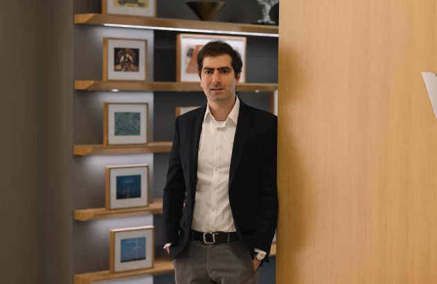 Homem de blazer preto e camisa branca, mãos nos bolsos, encostado em madeira. Prateleiras com quadros ao fundo.