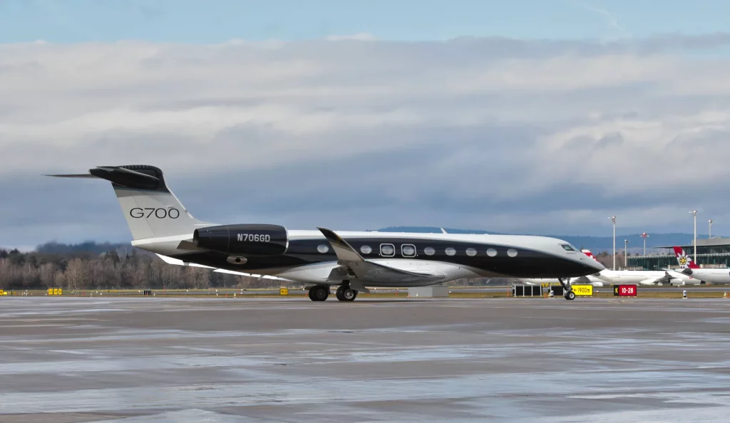 A imagem mostra um jato particular G700 estacionado em um aeroporto sob um céu nublado. O avião tem um design moderno em cores escuras, com "G700" na cauda e "N706GD" perto dos motores, sobre uma pista molhada que reflete a luz. Ao fundo, aparecem árvores, edifícios, outra aeronave e sinalização, sugerindo um ambiente típico e movimentado de aeroporto em dia nublado.