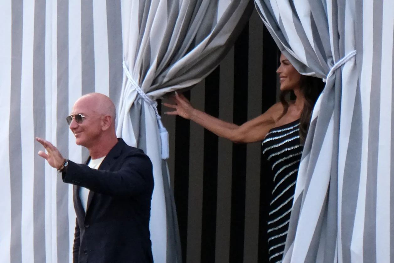A imagem mostra duas pessoas em um ambiente ao ar livre. À esquerda, um homem careca usando óculos de sol, terno escuro e camiseta clara sorri e acena, enquanto à direita, uma mulher com vestido preto listrado segura uma cortina, sorrindo para ele; ambos em um cenário elegante com cortinas listradas em branco e cinza.