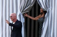 A imagem mostra duas pessoas em um ambiente ao ar livre. À esquerda, um homem careca usando óculos de sol, terno escuro e camiseta clara sorri e acena, enquanto à direita, uma mulher com vestido preto listrado segura uma cortina, sorrindo para ele; ambos em um cenário elegante com cortinas listradas em branco e cinza.