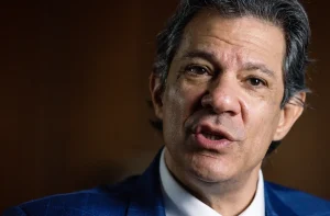 Homem em terno azul, com expressão séria e boca aberta, como se estivesse falando. Fundo escuro e desfocado.