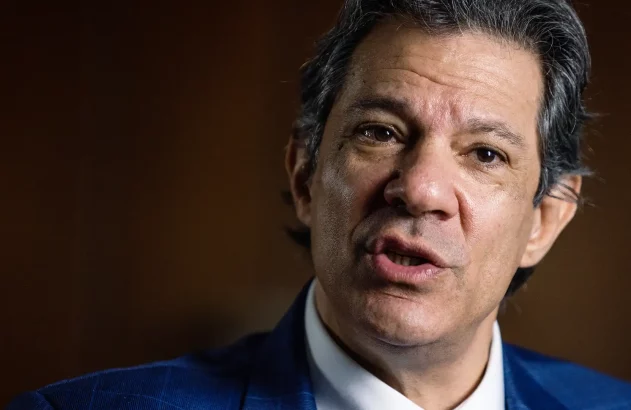 Homem em terno azul, com expressão séria e boca aberta, como se estivesse falando. Fundo escuro e desfocado.
