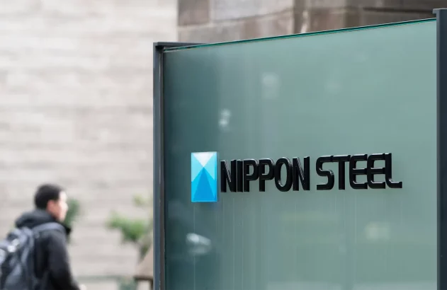 Placa de vidro com logotipo e nome "NIPPON STEEL" em letras pretas e estilo moderno. Logotipo azul e tridimensional à esquerda, fundo desfocado com parede de tijolos cinza e homem de casaco escuro com mochila.