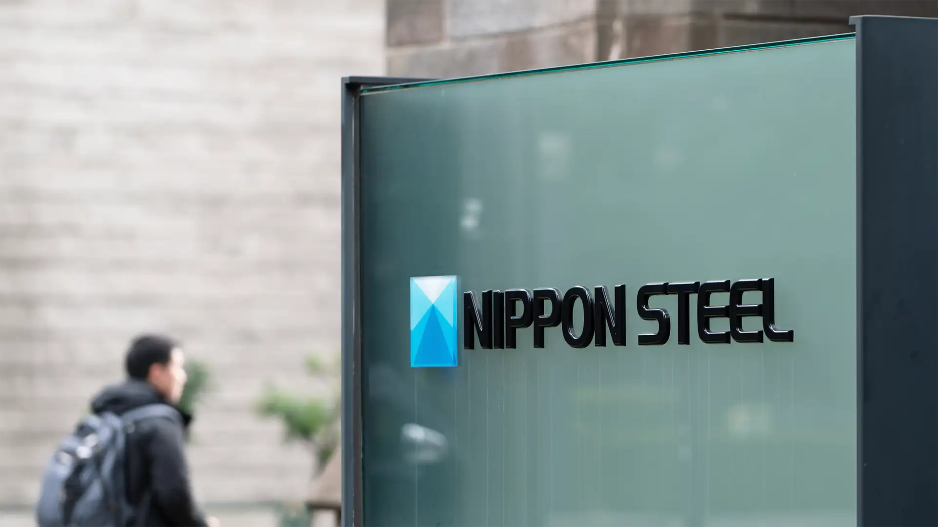 Placa de vidro com logotipo e nome "NIPPON STEEL" em letras pretas e estilo moderno. Logotipo azul e tridimensional à esquerda, fundo desfocado com parede de tijolos cinza e homem de casaco escuro com mochila.