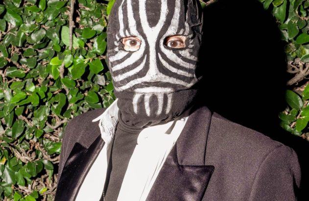 Pessoa com máscara estilo zebra, olhos visíveis, terno preto, camisa branca, colar, fundo verde de hera.