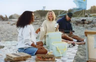 Três pessoas fazendo piquenique com pizza na praia. Torre de salva-vidas ao fundo.