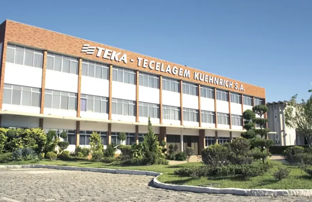 Edifício da TEKA - TECELAGEM KUEHNRICH S.A., com paisagismo frontal e céu azul.