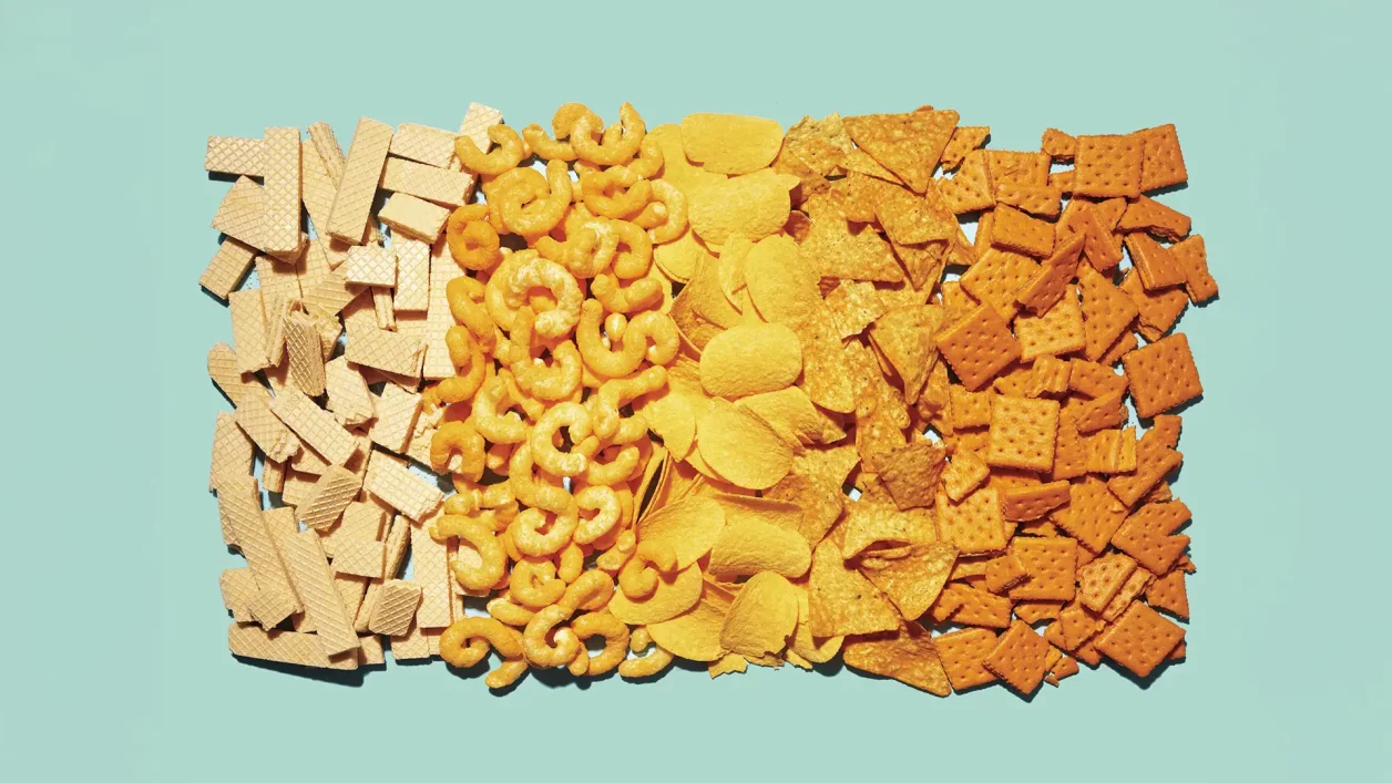 Variedade de salgadinhos organizados em faixas sobre fundo azul claro. Sequência de wafers claros, salgadinhos espiralados laranja, batatas fritas onduladas amarelas, nachos triangulares alaranjados e biscoitos quadrados laranja tostados.