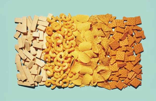 Variedade de salgadinhos organizados em faixas sobre fundo azul claro. Sequência de wafers claros, salgadinhos espiralados laranja, batatas fritas onduladas amarelas, nachos triangulares alaranjados e biscoitos quadrados laranja tostados.
