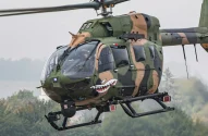 foto de um helicótero verde camuflado voando num céu cinza, com árvores ao longe