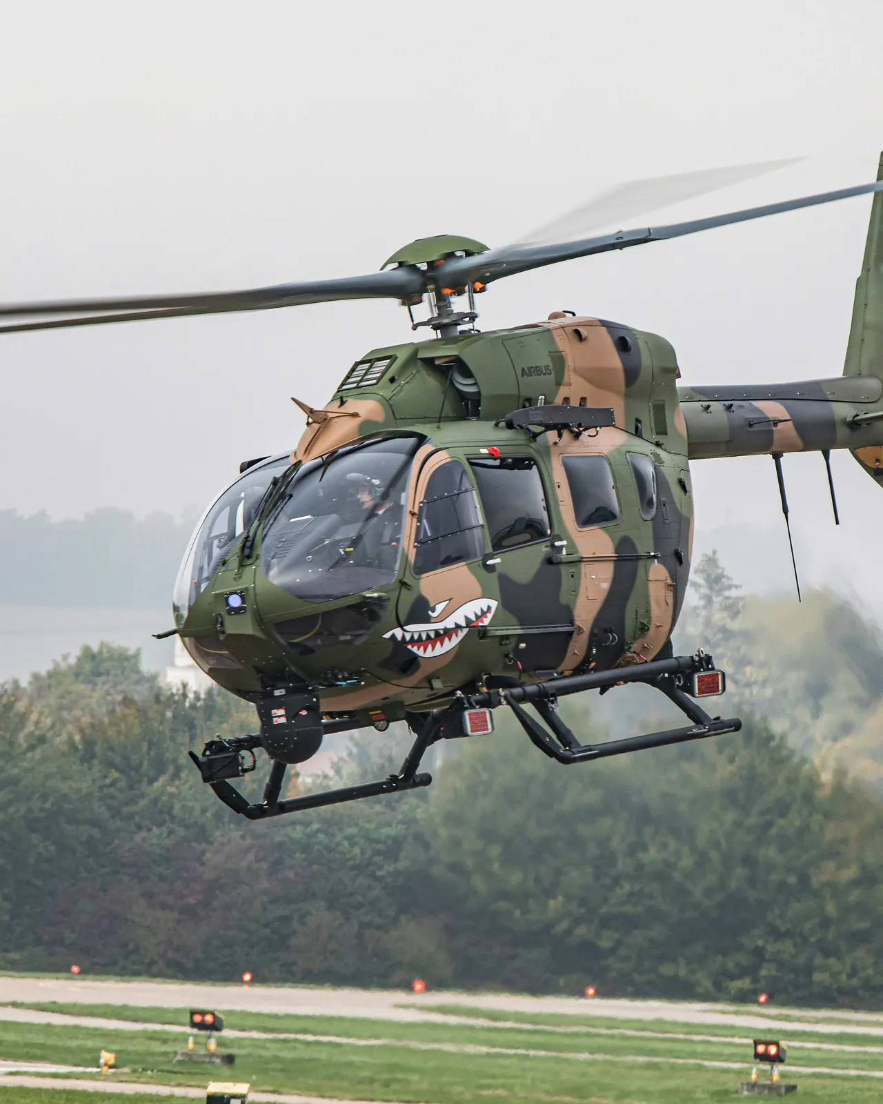 foto de um helicótero verde camuflado voando num céu cinza, com árvores ao longe