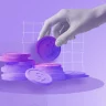 A imagem mostra um fundo roxo claro com textura granulada e uma pilha de moedas em roxo e rosa, destacando o símbolo de cifrão. Ao fundo, um gráfico de linhas e uma mão posicionando uma moeda no topo sugerem temas de investimento e economia.