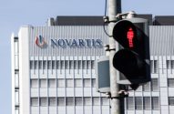 Foto do logotipo da Novartis no alto de um prédio cinza, à frente temos um semáforo para pedestre com o boneco vermelho em destaqu