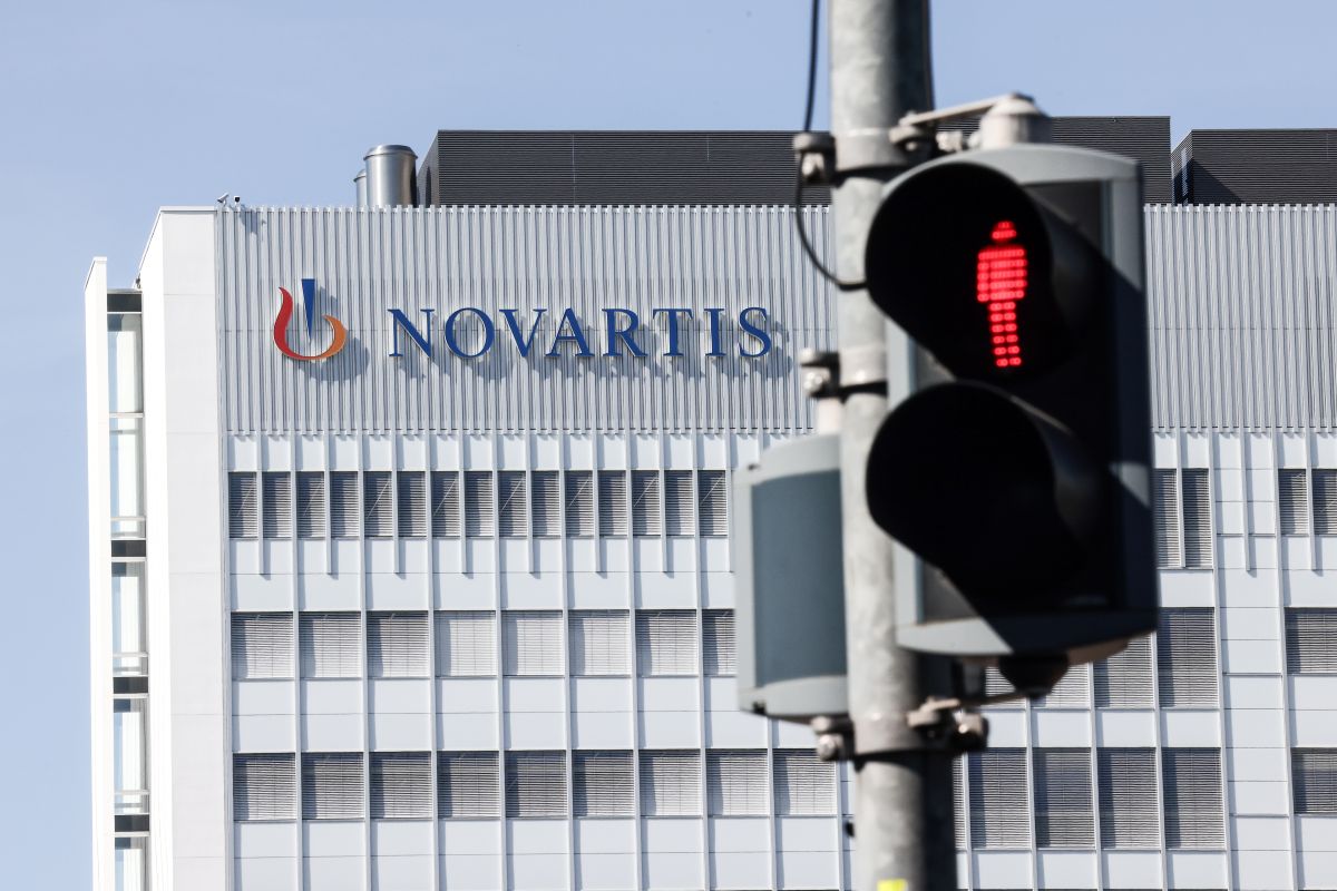 Foto do logotipo da Novartis no alto de um prédio cinza, à frente temos um semáforo para pedestre com o boneco vermelho em destaqu