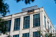 A imagem mostra a parte superior de um edifício comercial moderno com o logotipo da AWS na fachada, janelas de vidro azuladas e linhas retas. O logotipo inclui uma seta laranja em formato de sorriso. O céu está claro e galhos de árvores aparecem desfocados em primeiro plano.