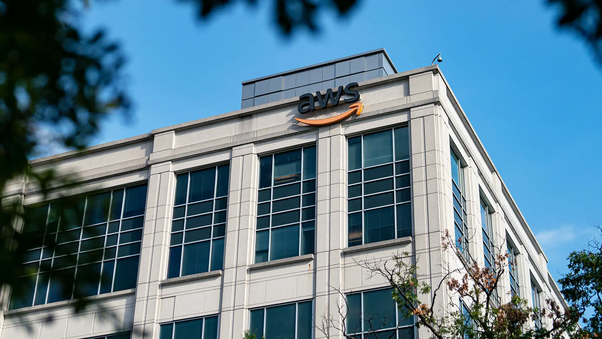 A imagem mostra a parte superior de um edifício comercial moderno com o logotipo da AWS na fachada, janelas de vidro azuladas e linhas retas. O logotipo inclui uma seta laranja em formato de sorriso. O céu está claro e galhos de árvores aparecem desfocados em primeiro plano.