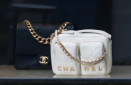 A imagem mostra duas bolsas de luxo. A bolsa à esquerda é preta com acolchoado em padrão de losangos e uma alça de corrente dourada, enquanto a bolsa à direita é branca, com duas bolsas frontais com zíperes dourados e o nome CHANEL entre elas, além de uma alça de corrente dourada com seção de couro branco.