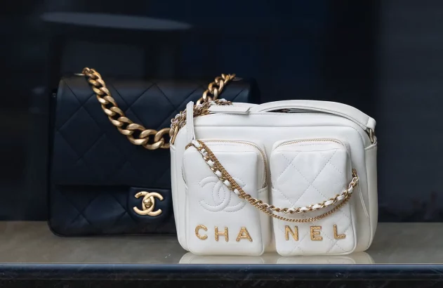 A imagem mostra duas bolsas de luxo. A bolsa à esquerda é preta com acolchoado em padrão de losangos e uma alça de corrente dourada, enquanto a bolsa à direita é branca, com duas bolsas frontais com zíperes dourados e o nome CHANEL entre elas, além de uma alça de corrente dourada com seção de couro branco.
