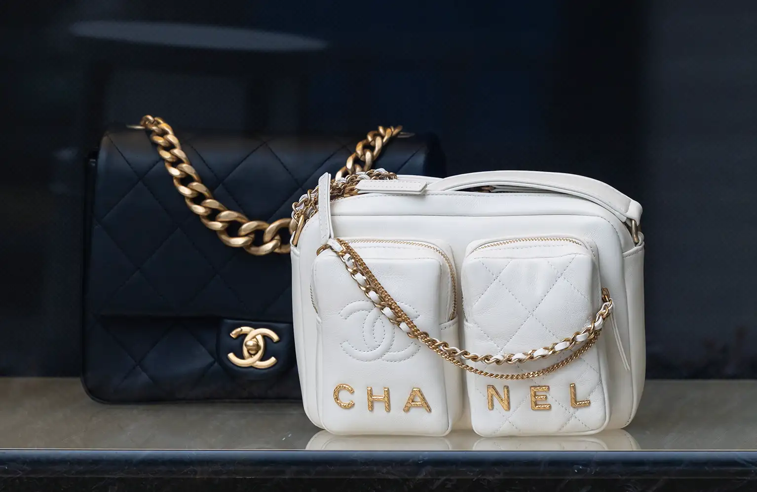 A imagem mostra duas bolsas de luxo. A bolsa à esquerda é preta com acolchoado em padrão de losangos e uma alça de corrente dourada, enquanto a bolsa à direita é branca, com duas bolsas frontais com zíperes dourados e o nome CHANEL entre elas, além de uma alça de corrente dourada com seção de couro branco.