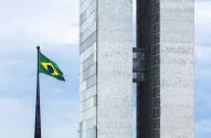 A imagem mostra a fachada do Congresso Nacional do Brasil, com suas duas torres gêmeas de mármore branco, passarelas interligadas e um céu azul claro ao fundo. À esquerda, a bandeira brasileira está hasteada em um mastro preto.