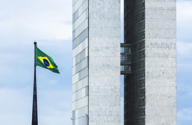 A imagem mostra a fachada do Congresso Nacional do Brasil, com suas duas torres gêmeas de mármore branco, passarelas interligadas e um céu azul claro ao fundo. À esquerda, a bandeira brasileira está hasteada em um mastro preto.