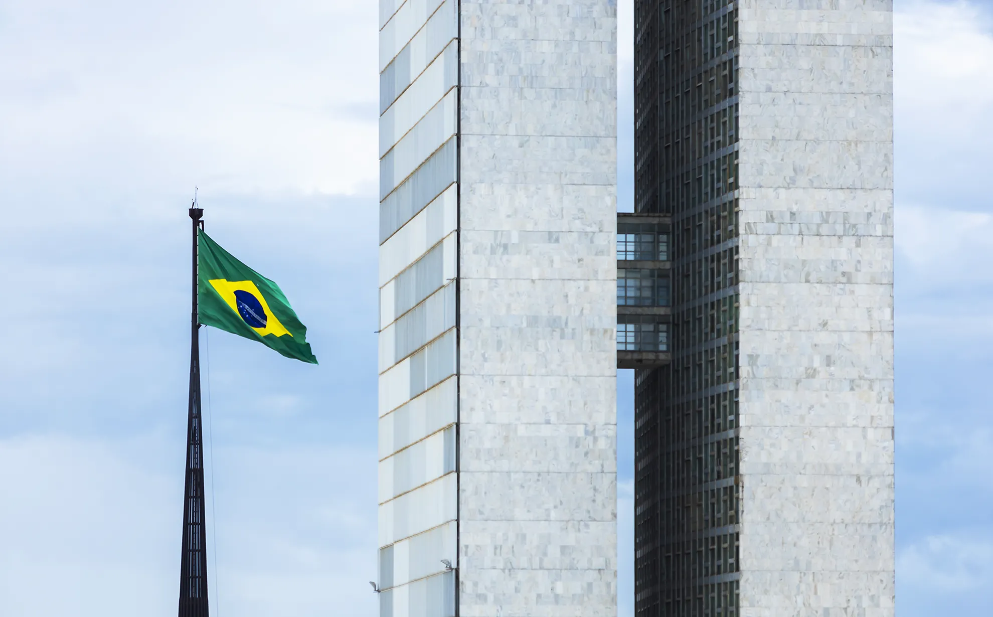 A imagem mostra a fachada do Congresso Nacional do Brasil, com suas duas torres gêmeas de mármore branco, passarelas interligadas e um céu azul claro ao fundo. À esquerda, a bandeira brasileira está hasteada em um mastro preto.