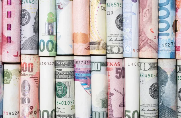 A imagem apresenta diversas notas de diferentes países, enroladas verticalmente e alinhadas lado a lado, exibindo uma colagem colorida que destaca a diversidade monetária global, com variações em valores, ícones e estilo artístico.