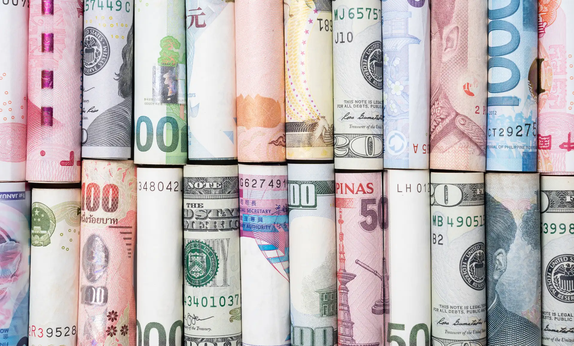 A imagem apresenta diversas notas de diferentes países, enroladas verticalmente e alinhadas lado a lado, exibindo uma colagem colorida que destaca a diversidade monetária global, com variações em valores, ícones e estilo artístico.