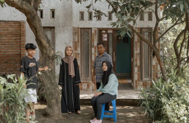 Um casal e um adolescente estão em pe embaixo de uma árvoore, e a filha adolescente está sentada em um banquinho na frente deles