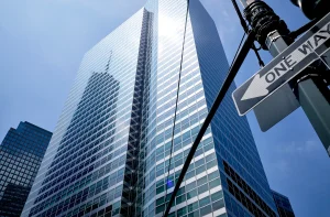 A imagem mostra um arranha-céu moderno com numerosas janelas espelhadas refletindo o céu azul. O edifício alto possui linhas retas e design contemporâneo. À direita, há um poste de luz com um sinal de "one way", indicando uma área urbana movimentada.