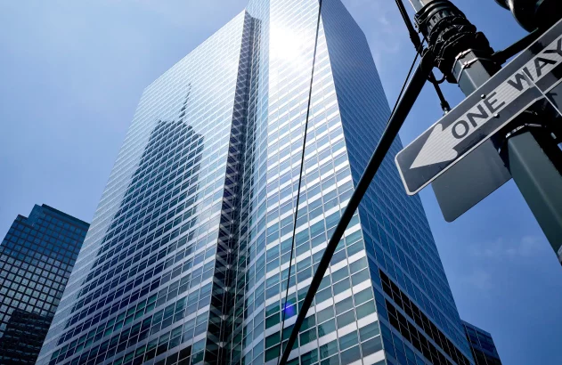 A imagem mostra um arranha-céu moderno com numerosas janelas espelhadas refletindo o céu azul. O edifício alto possui linhas retas e design contemporâneo. À direita, há um poste de luz com um sinal de "one way", indicando uma área urbana movimentada.
