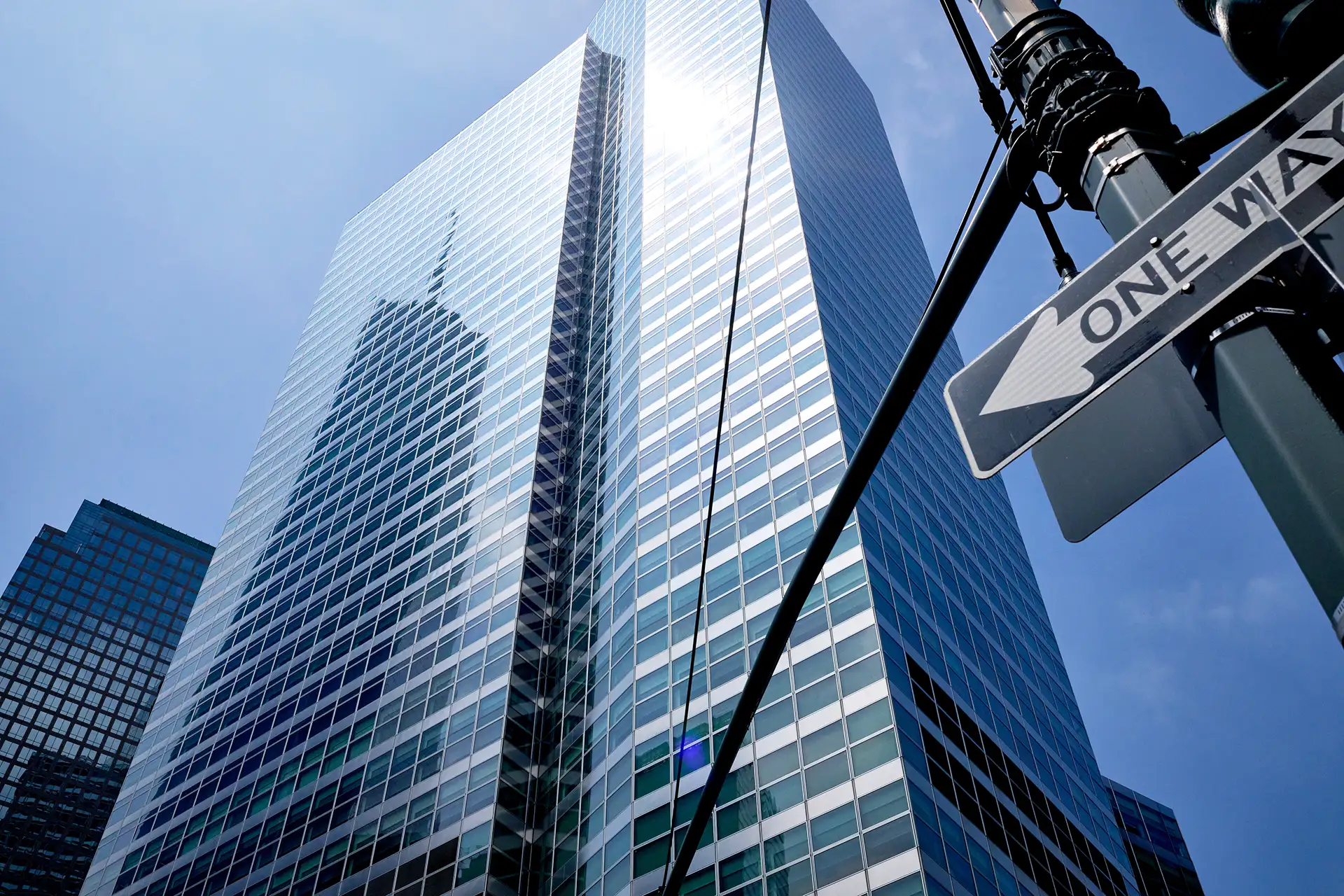 A imagem mostra um arranha-céu moderno com numerosas janelas espelhadas refletindo o céu azul. O edifício alto possui linhas retas e design contemporâneo. À direita, há um poste de luz com um sinal de "one way", indicando uma área urbana movimentada.