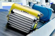 A imagem mostra três malas em um carrossel de bagagens no aeroporto: uma prata com listras e rodas, uma amarela similar e uma azul parcialmente visível. O carrossel metálico está em movimento, com etiquetas de bagagem nas alças das malas, em um ambiente de aeroporto movimentado.