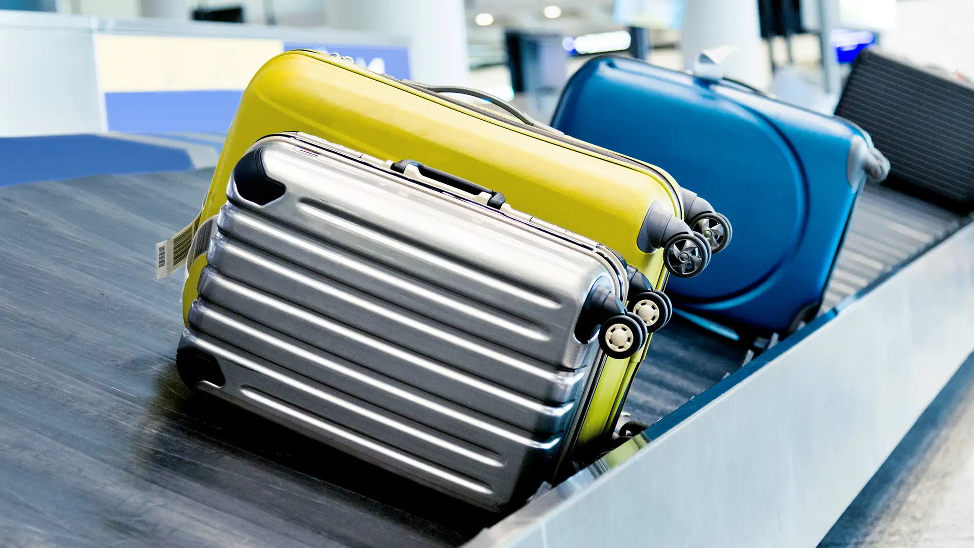 A imagem mostra três malas em um carrossel de bagagens no aeroporto: uma prata com listras e rodas, uma amarela similar e uma azul parcialmente visível. O carrossel metálico está em movimento, com etiquetas de bagagem nas alças das malas, em um ambiente de aeroporto movimentado.