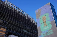 A imagem apresenta o estádio MetLife à esquerda, com estrutura metálica cinza e vidro, e o nome em letras grandes na fachada. À direita, um painel exibe uma taça dourada com "2026" em verde e azul, e "FIFA" relacionada à Copa do Mundo de 2026, acompanhada de "NEW YORK" e "NEW JERSEY".