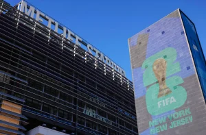 A imagem apresenta o estádio MetLife à esquerda, com estrutura metálica cinza e vidro, e o nome em letras grandes na fachada. À direita, um painel exibe uma taça dourada com "2026" em verde e azul, e "FIFA" relacionada à Copa do Mundo de 2026, acompanhada de "NEW YORK" e "NEW JERSEY".