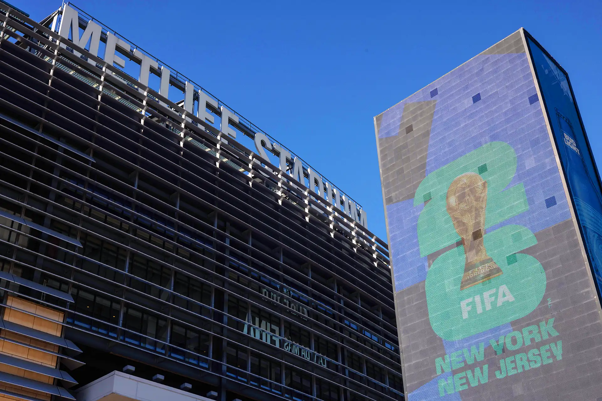 A imagem apresenta o estádio MetLife à esquerda, com estrutura metálica cinza e vidro, e o nome em letras grandes na fachada. À direita, um painel exibe uma taça dourada com "2026" em verde e azul, e "FIFA" relacionada à Copa do Mundo de 2026, acompanhada de "NEW YORK" e "NEW JERSEY".